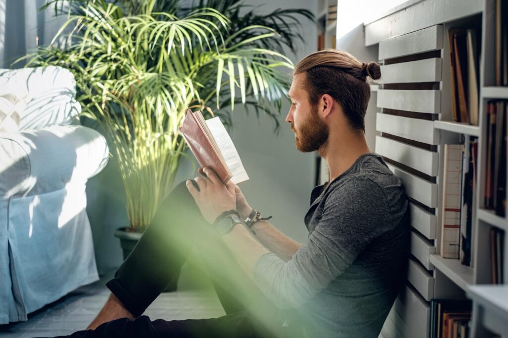 A bearded man is reading a book in a room with green plants