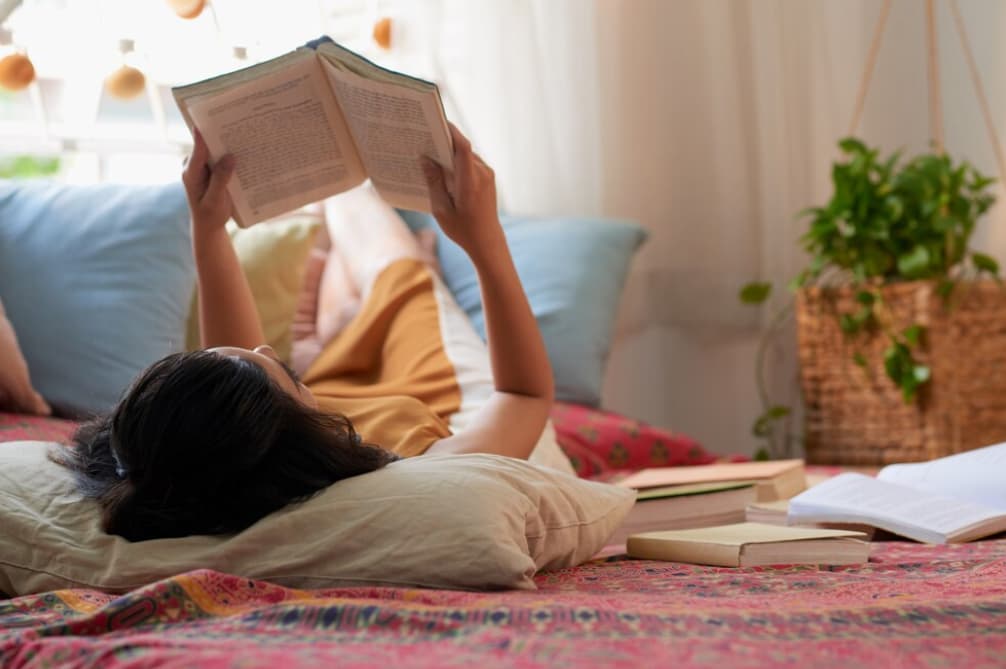 A person lies on a bed reading a book with a cozy room backdrop